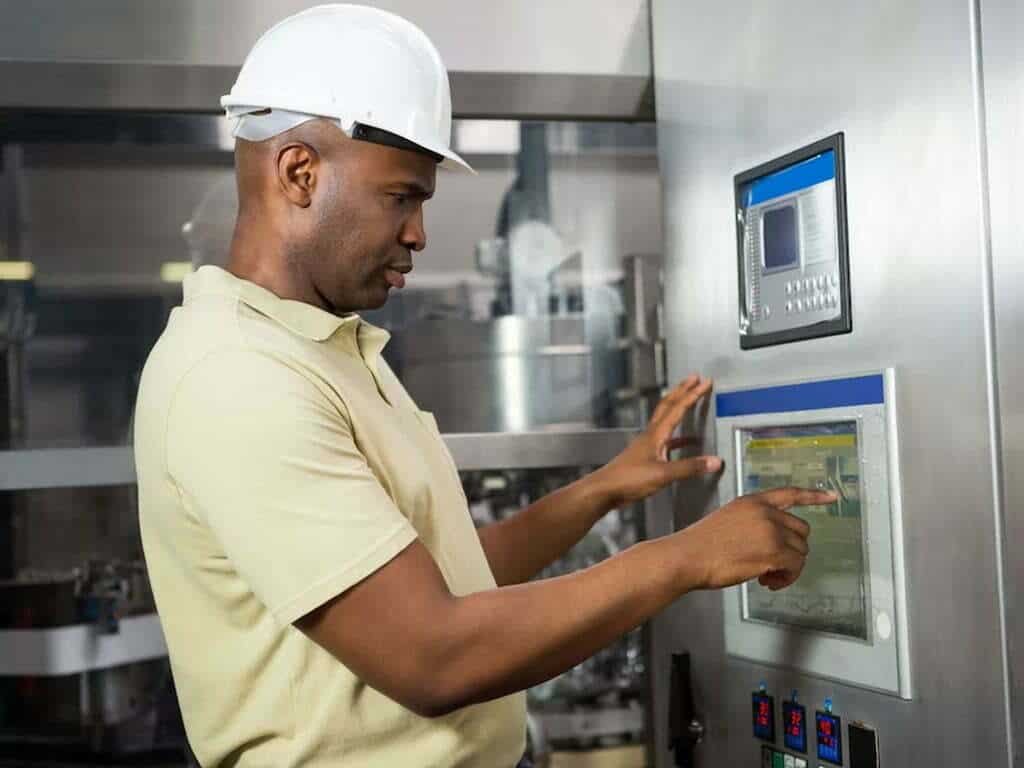 A factory worker wearing a hard hat is operating a touch screen control panel on a machine.