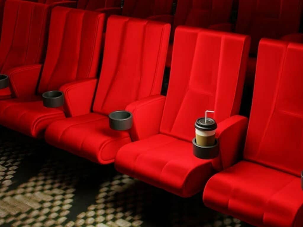 Rows of empty, plush red seats with drink holders are shown in a movie theater. One cup holds a drink with a straw.