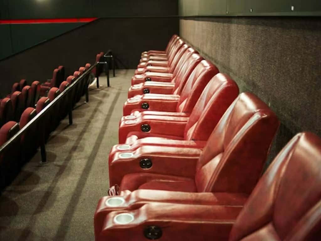 Rows of empty, reclining red leather seats with cup holders are shown in a movie theater.