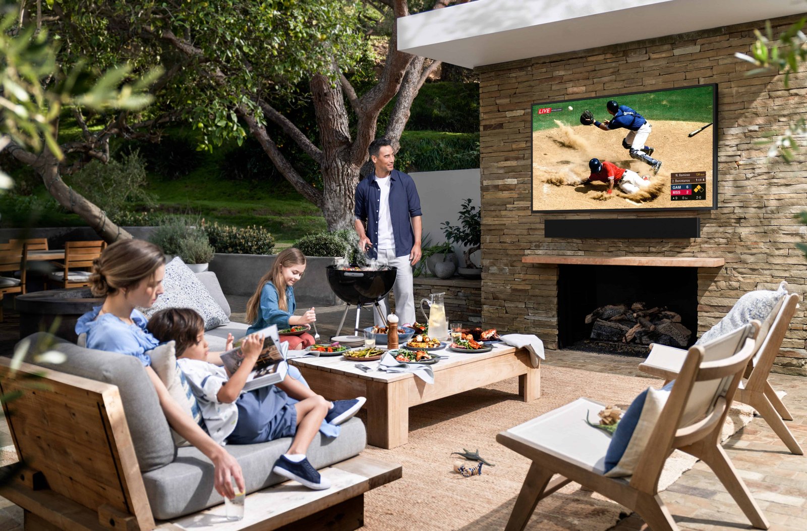 A family relaxes on a patio with a TV showing a baseball game.