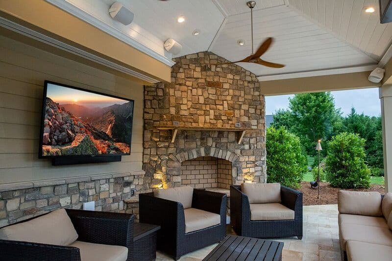 A luxurious outdoor patio features a stone fireplace, a mounted TV, and comfortable wicker furniture.