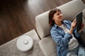 Woman on couch with tablet, robot vacuum on floor.