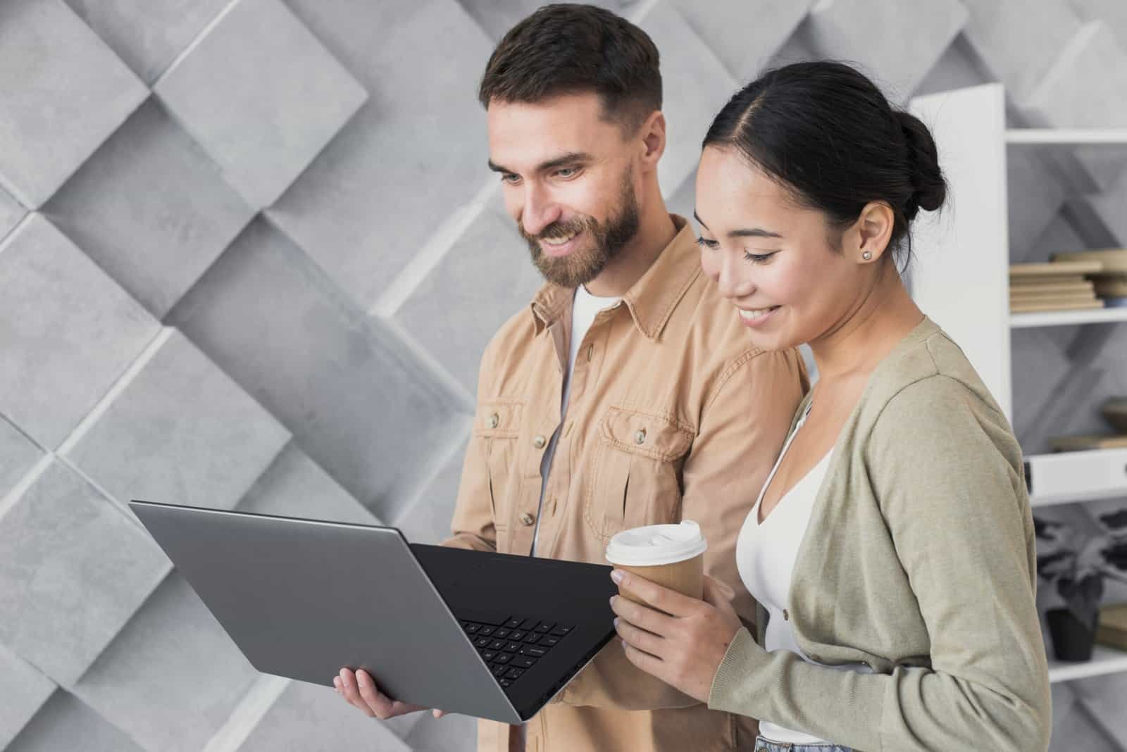Two smiling people looking at a laptop, one holding coffee.