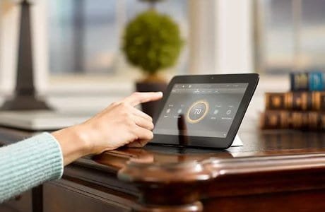 A person is using a tablet to control a smart thermostat, which is displaying the temperature as 70 degrees.