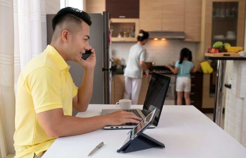 Man working on laptop and phone, family in background.